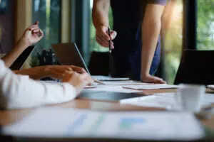 image of a man pointing a pencil at sheets of paper on an office desk surrounded by colleagues
